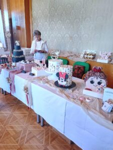 Ein Tisch voller kunstvoll dekorierter Kuchen auf einer Backmesse, eine Frau betrachtet ihr Handy. Einblick in regionale Backkultur.