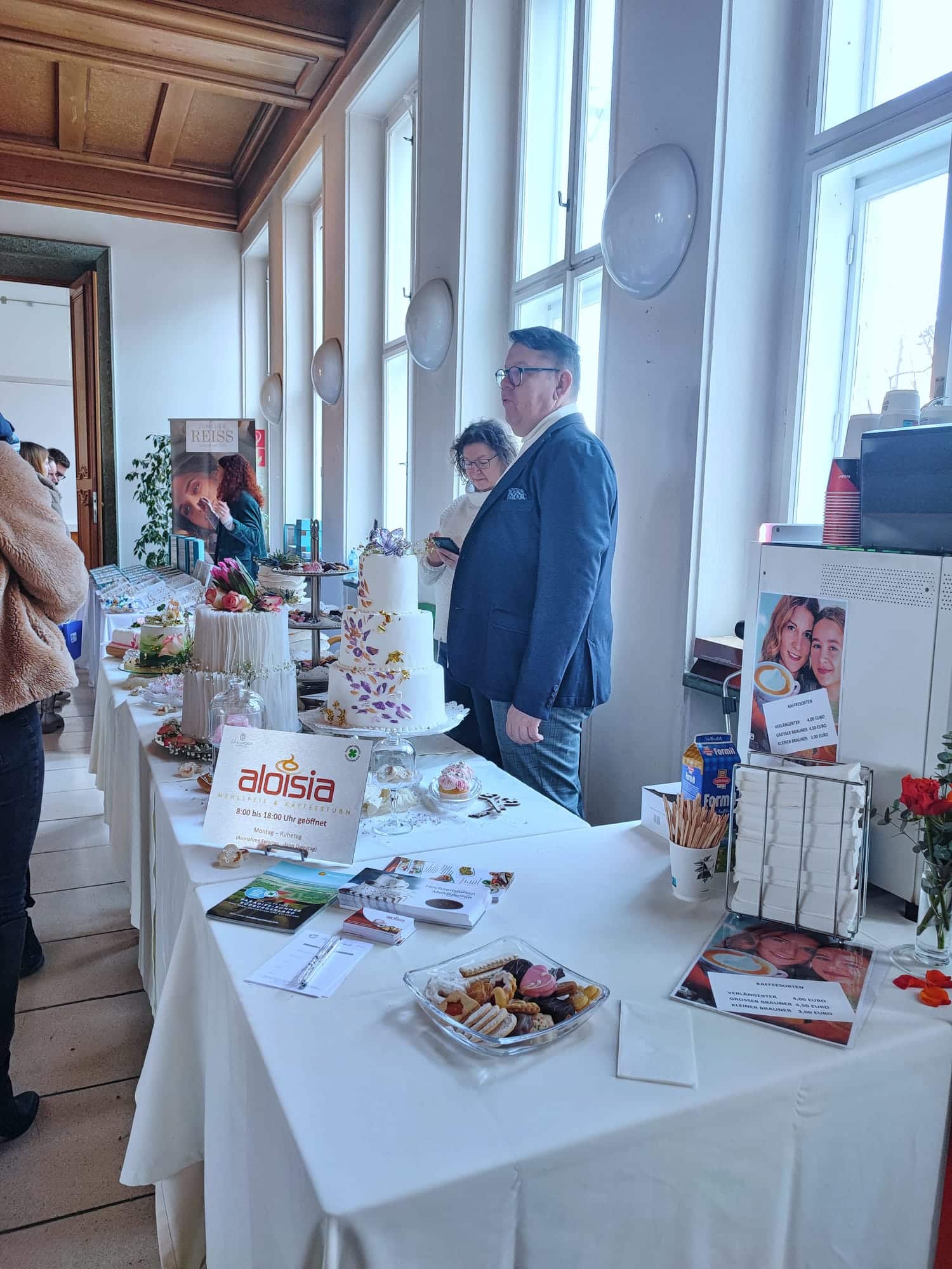 Messeaufbau mit liebevoll dekoriertem Stand, Kuchen und Prospekten; Personen im Gespräch. Passt zu Prima Magazins regionalem Flair.