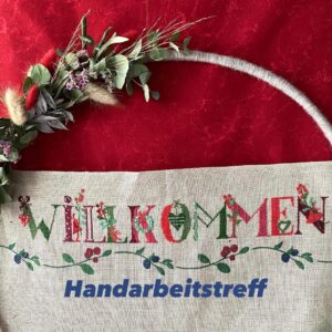 Besticktes Willkommen-Schild mit Blumenmuster über einem roten Stoff, lädt zum Handarbeitstreff im regionalen Magazin ein.