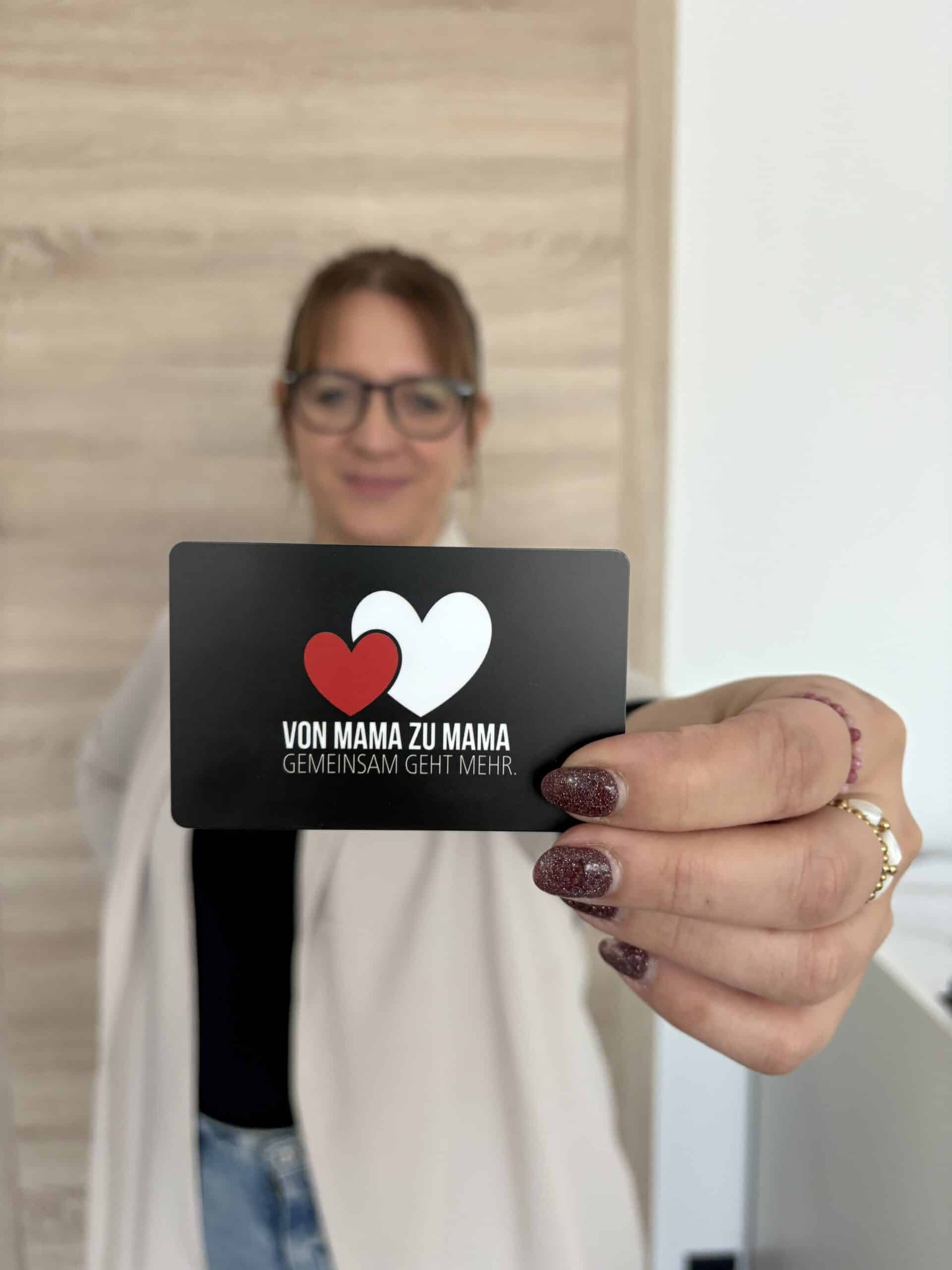 Eine Frau hält eine Karte mit zwei Herzen und dem Text Von Mama zu Mama, Gemeinsam geht mehr in die Kamera.