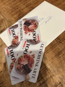 Drei SPAR-Geschenkkarten im Wert von je 100 Euro liegen auf einem Holztisch, neben einem Brief mit handschriftlicher Notiz.