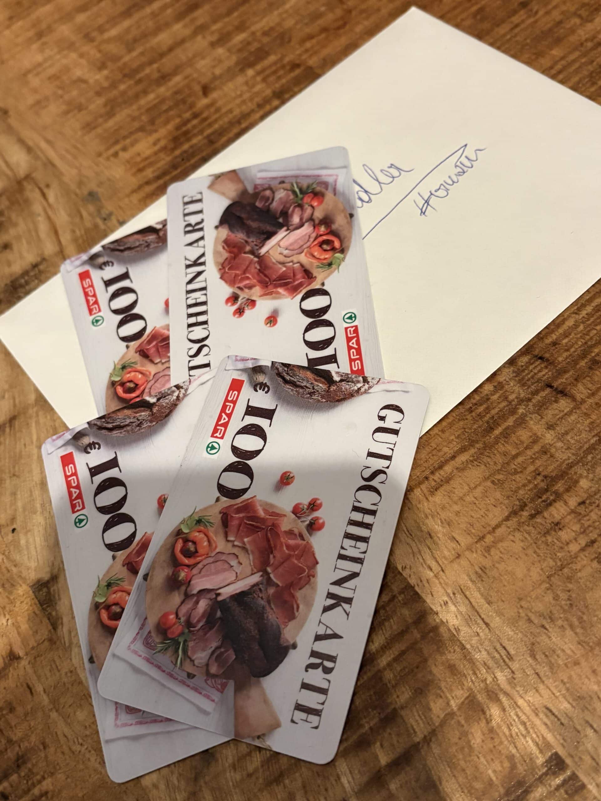 Drei SPAR-Geschenkkarten im Wert von je 100 Euro liegen auf einem Holztisch, neben einem Brief mit handschriftlicher Notiz.