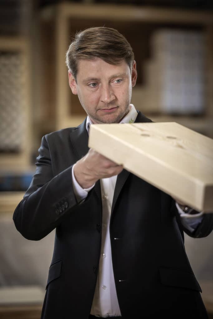 Michael Oberfeichtner©OK Haus Ein Mann im Anzug bewertet konzentriert ein Holzbrett in einer Werkstattumgebung.