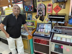 Ein Mann lächelt in einer Sammlung nostalgischer Jukeboxen und Musikautomaten, präsentiert in einem einladenden, bunten Raum.