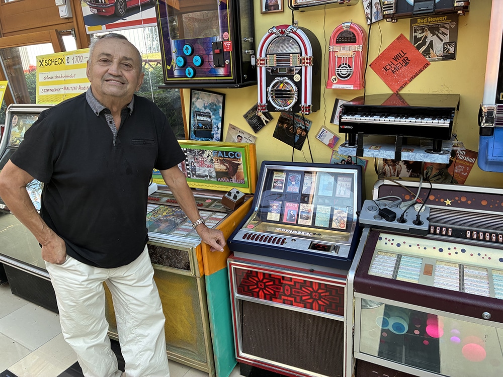 Ein Mann lächelt in einer Sammlung nostalgischer Jukeboxen und Musikautomaten, präsentiert in einem einladenden, bunten Raum.