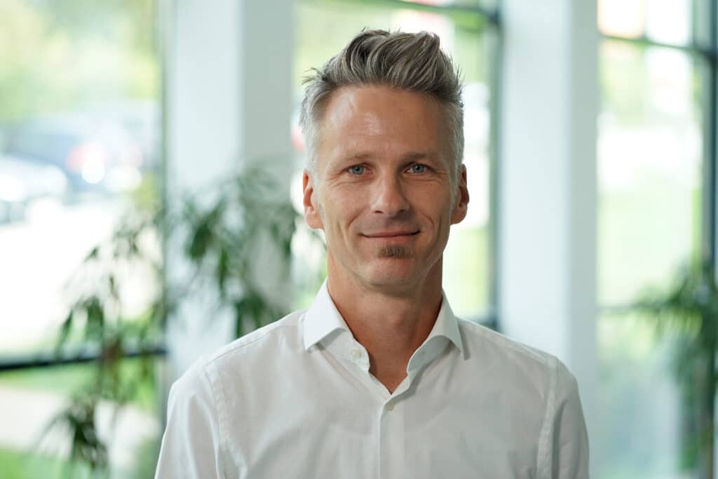 Porträt eines lächelnden Mannes in weißem Hemd vor einem hellen, pflanzenreichen Hintergrund.