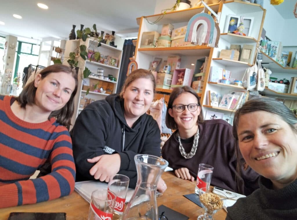Wiederwertvoll_HBLA©WiederWertvoll Vier Frauen lächeln an einem Tisch in einem gemütlichen Café, umgeben von Regalen voller Bücher und Dekorationen.