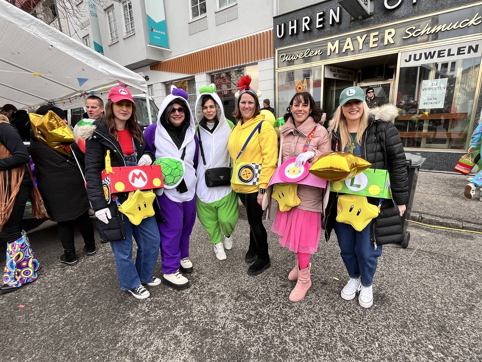 Menschen in bunten Kostümen feiern Karneval auf einer belebten Straße, vor einem Geschäft mit regionalem Flair, ideal für Prima Magazin.