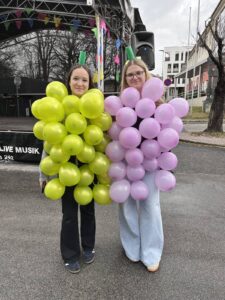 Zwei Personen in bunten Traubenkostümen stehen lächelnd vor einer Bühne auf einem regionalen Straßenfest.