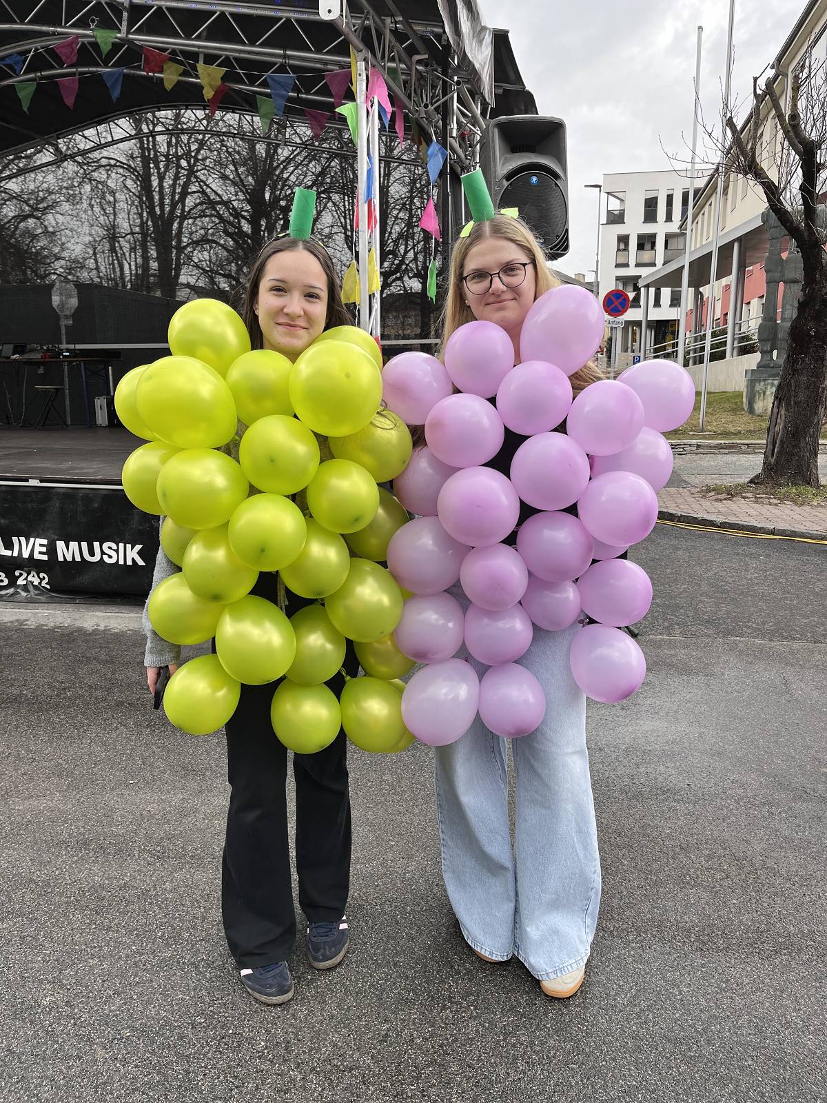 Zwei Personen in bunten Traubenkostümen stehen lächelnd vor einer Bühne auf einem regionalen Straßenfest.