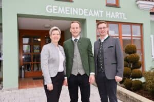 Drei Personen in traditionellen Trachten in Hartl vor dem Gemeindeamt, repräsentieren lokale Kultur für Prima Magazin.