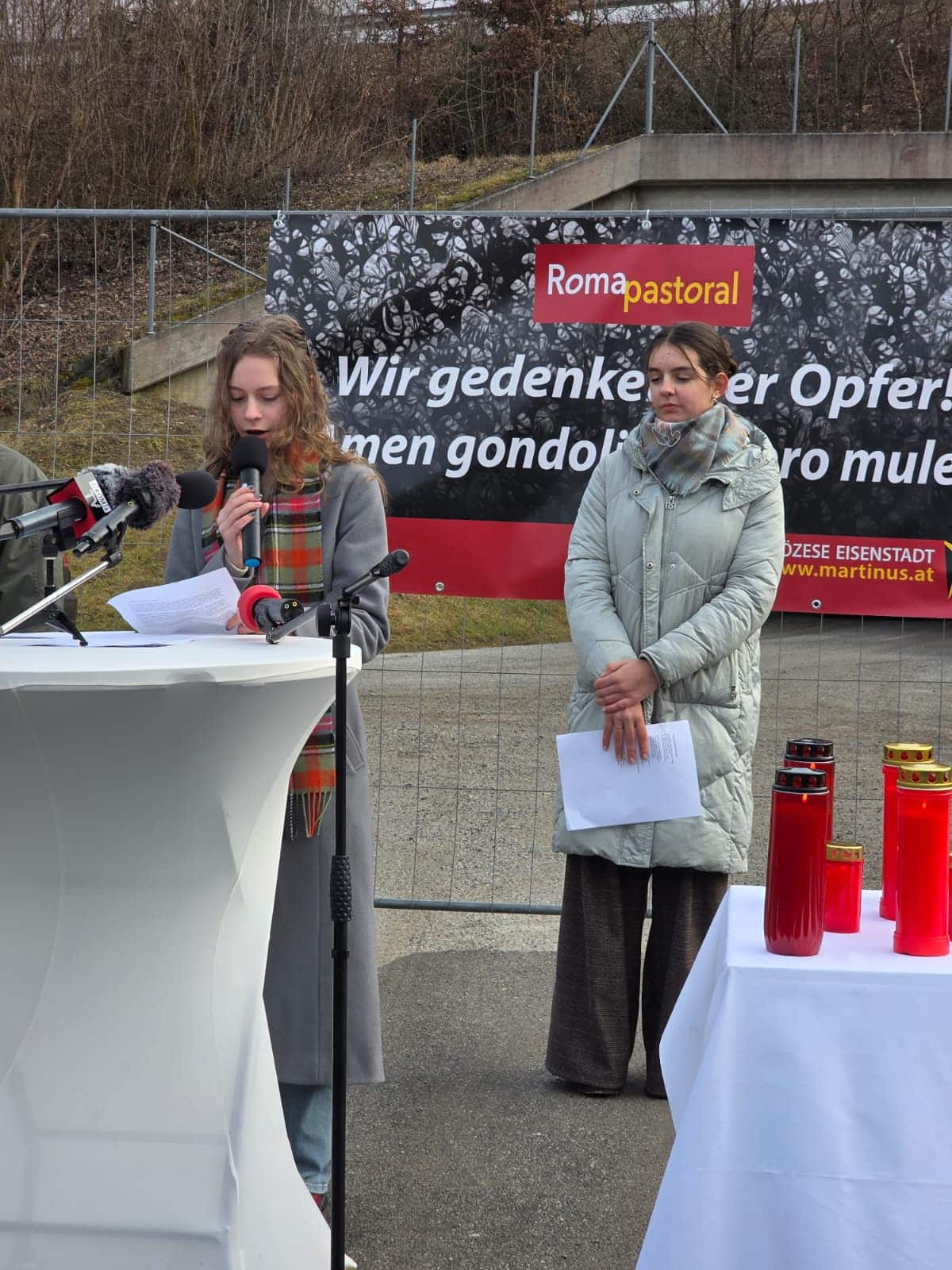 Gedenkveranstaltung im Freien mit zwei Frauen am Mikrofon vor einem Banner von Roma Pastoral, rote Kerzen im Vordergrund.