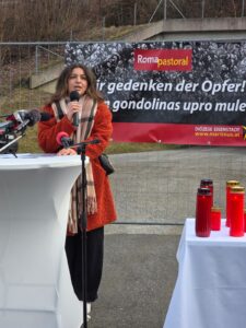 Eine Frau hält bei einer Gedenkveranstaltung für Opfer eine Rede, vor einem Banner mit Kerzen auf einem Tisch.
