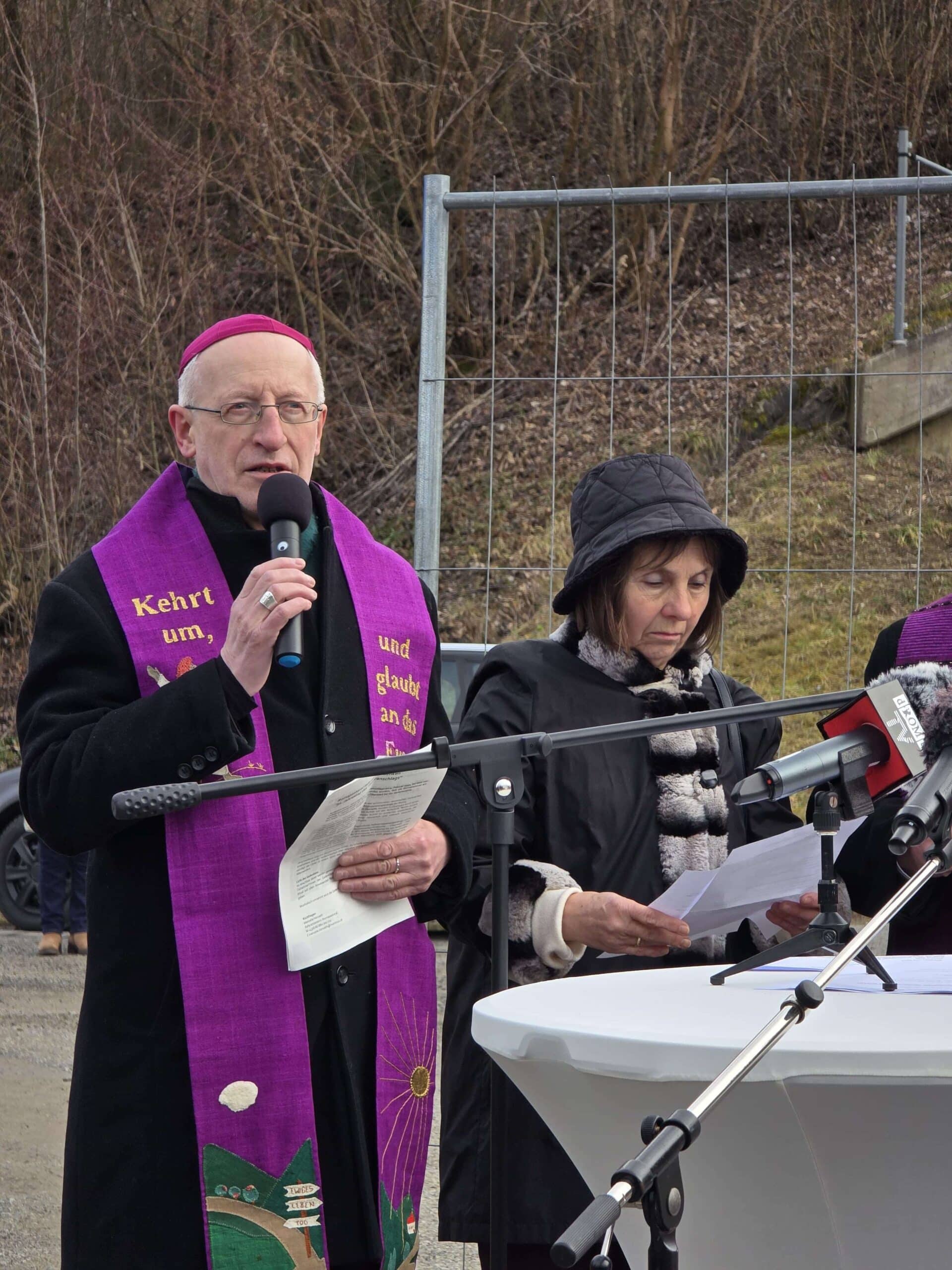 Ein älterer Mann spricht bei einer Veranstaltung draußen, begleitet von einer Frau mit Papier in der Hand, vor Mikrofonen.