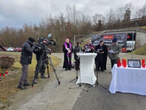 Gedenkveranstaltung im Freien mit Geistlichen und Presse, mit Kerzen und Foto auf Tisch, gefilmt von Kamerateam für regionales Magazin.