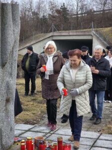 Ältere Menschen bei Gedenkveranstaltung, halten rote Kerzen in Händen, erinnern an besondere Momente, im Freien vor Brücke.