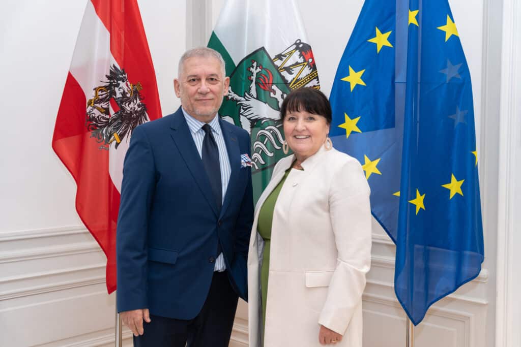 Zwei Personen posieren vor österreichischer, steirischer und EU-Flagge, symbolisch für internationale Vernetzung und Zusammenarbeit.